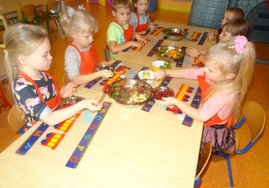 Grupa dzieci siedzi przy stole Trzy dziewczynki trzymają w ręku łyżki i przekładają owoce do dużej miski z pokrojonymi owocami.